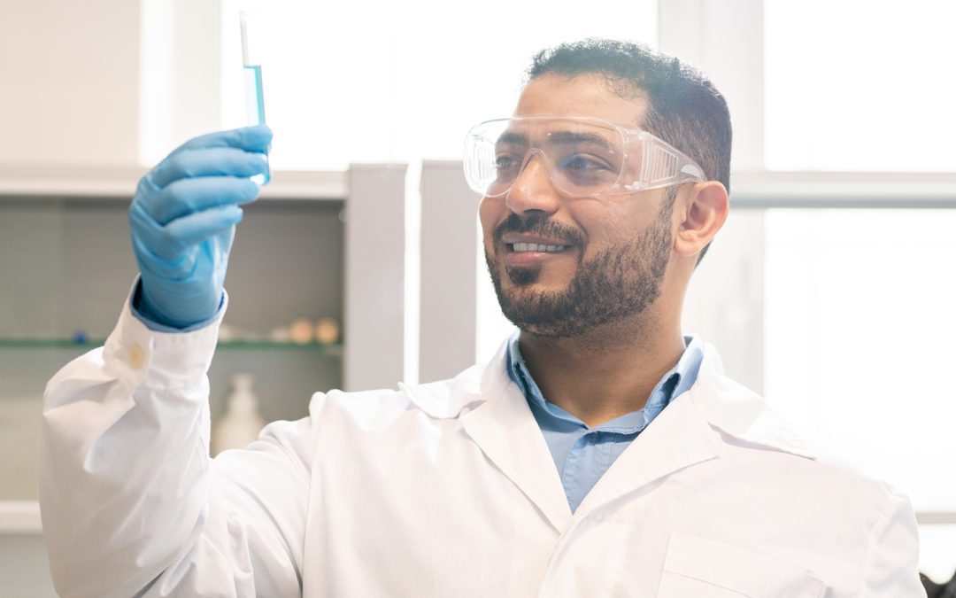 Positive satisfied young middle eastern scientist with beard wearing rubber gloves and safety goggles holding test tube with liquid while inventing medication in laboratory
