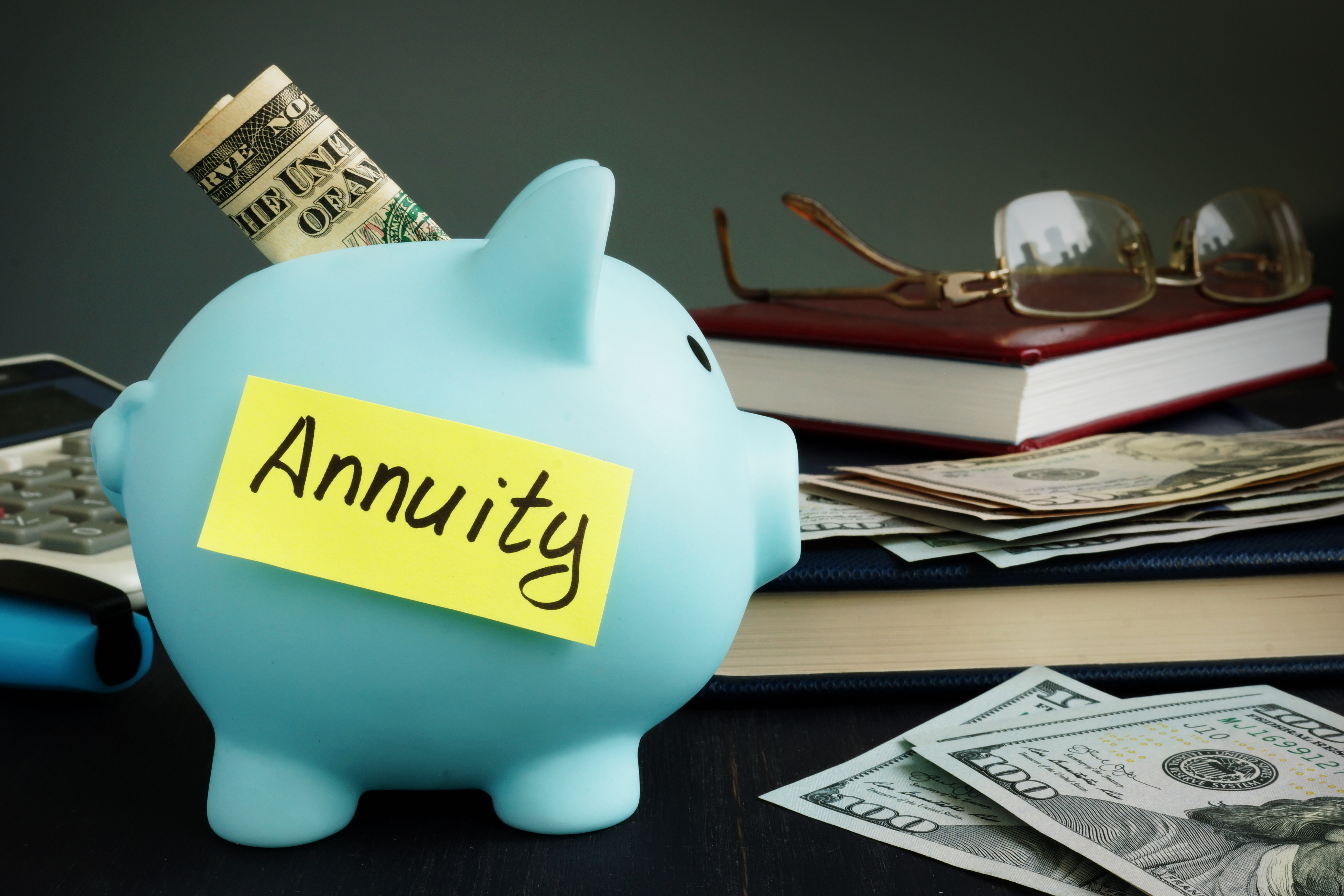 Annuity written on a yellow post-it and stuck to a blue piggy bank sitting in front of stack of books